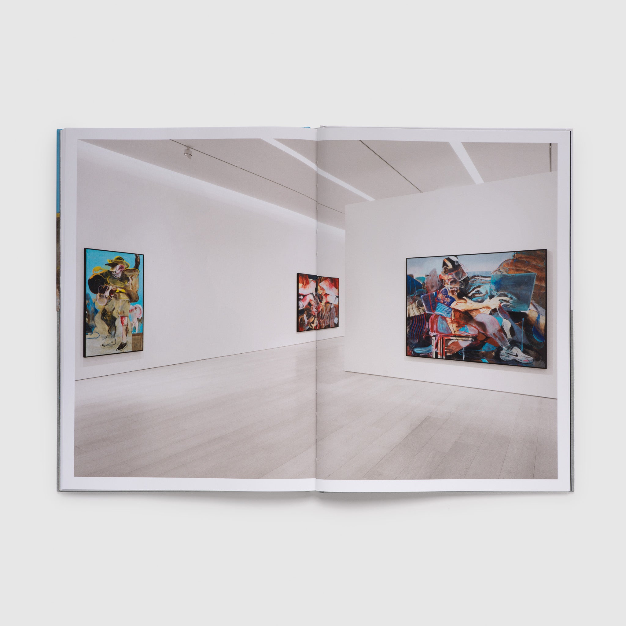 The book is open to two pages with a photograph of the exhibition across both. There are three paintings hanging on white walls.