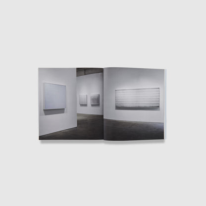 The book is open to two pages. One image spans both pages, it is of the Donovan exhibition in a white gallery with cement floors. The Donovan works are hanging on the walls of the gallery.