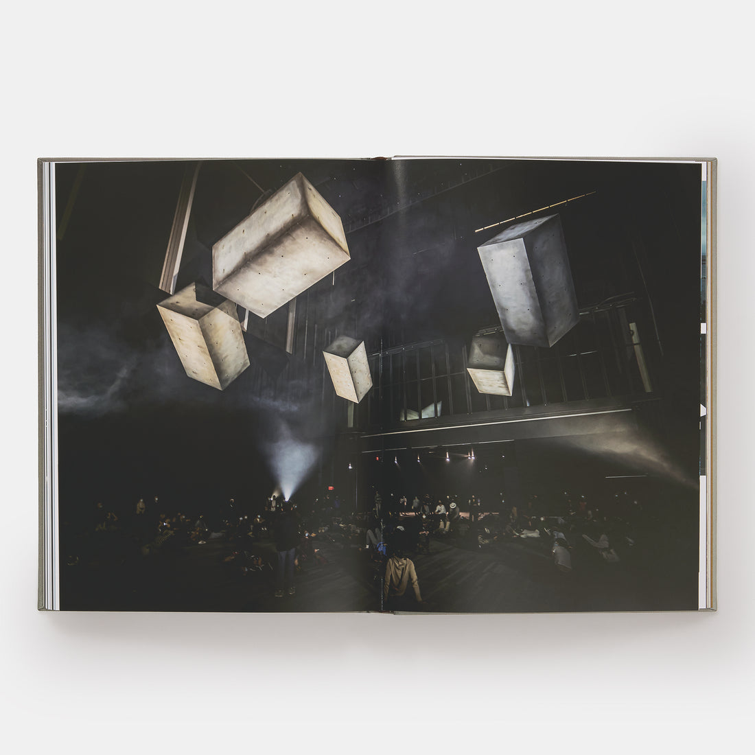 The book is open to two pages with an image of Drift works in a gallery across both pages. There are large blocks suspended from the ceiling and a crowd of people below looking up at them.