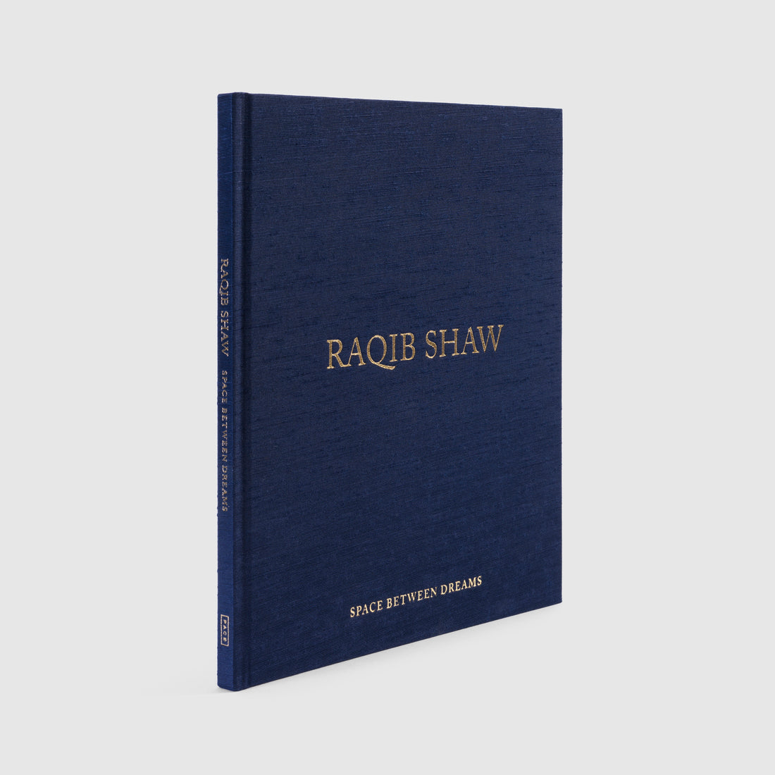 Fabric blue book with gold text that reads "Raqib Shaw" on a light gray background